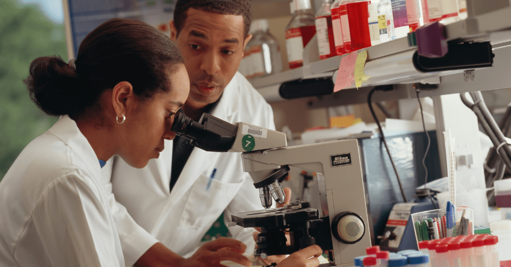 A female looks through a microscope while a male companion looks on in the lab.