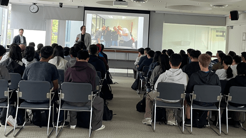 Students in an audience, two presenters and a slide show 