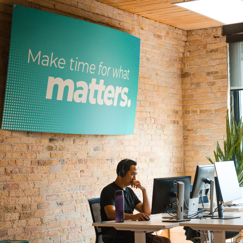 Loopio employee sitting infront of a desktop with a huge sign that reads 'Make time for what matters'