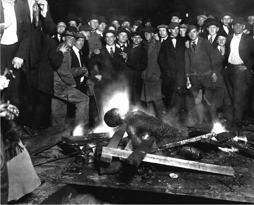 A mob of white men gathers around a burning African-American individual.
