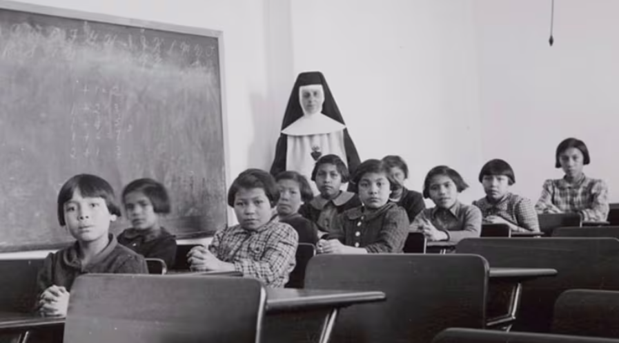 A group of female students and a nun pose in a classroom at Cross Lake Indian Residential School in Cross Lake, Manitoba, February 1940