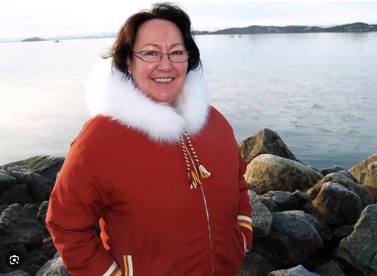Person in orange fur-trimmed jacket standing on rocky shoreline with water and distant land in the background