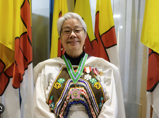 Person in traditional attire with decorations and medals; face blurred; multiple yellow flags with red and black designs in the background