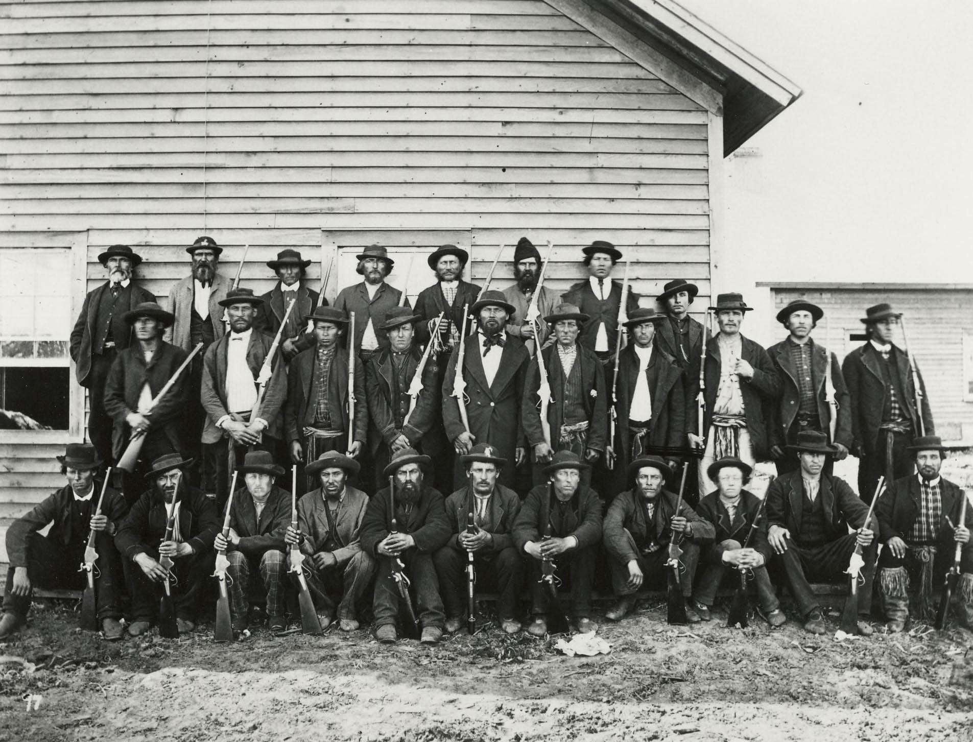 Group of 28 men posing in three rows in front of a wooden building, most holding rifles or shotguns and wearing hats and suits