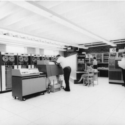 Peering into the 1403 printer is Richard Shirley. Marian Rankin is seated at centre in front of the IBM 7040. Sandra Hope is at rear. Mike Doyle, reading a sheet of printout, stands in front of the IBM 1401.