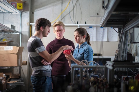 Three people stand in a lab looking at a red object for quantum research