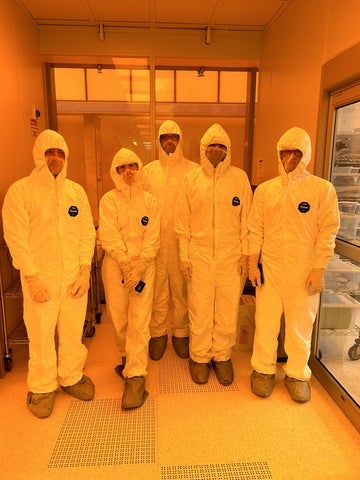 Five USEQIP students dressed in white cleanroom suits, standing in orange light