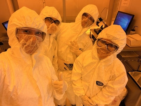 Four USEQIP students posing together in the clean room. They are all wearing white full-body suits and safety glasses.