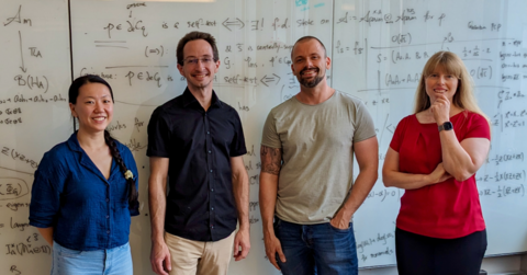 Four researchers speaking and listening to each other in front of a white board with math forumlas on it