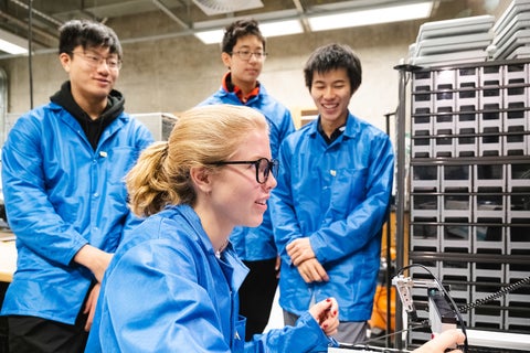 Three QSYS participants in blue lab coats watching a fourth participant who is controlling a quantum experiment