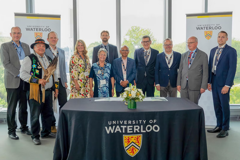 11 people standing by a University of Waterloo marked chair in front of a large window