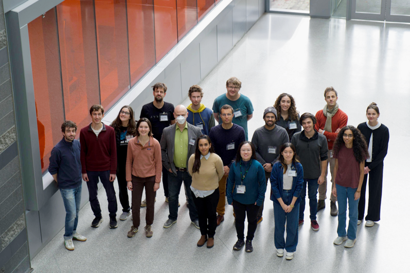 A group of 18 people standing in front of an orange wall in a big hallway looking up