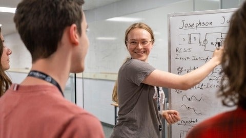 Woman writes physics formula on whitboard and looks at group of students smiling