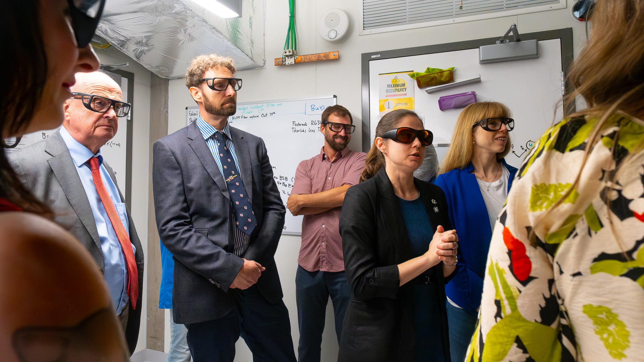 seven people standing in a bright room wearing protective glasses listening to a scientist speak