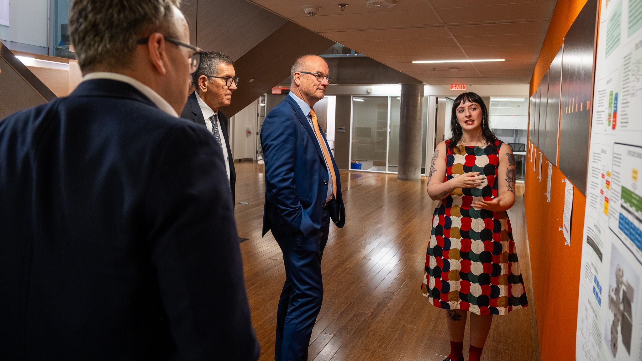 A woman on the right speaks to three men on the left in front of a poster wall
