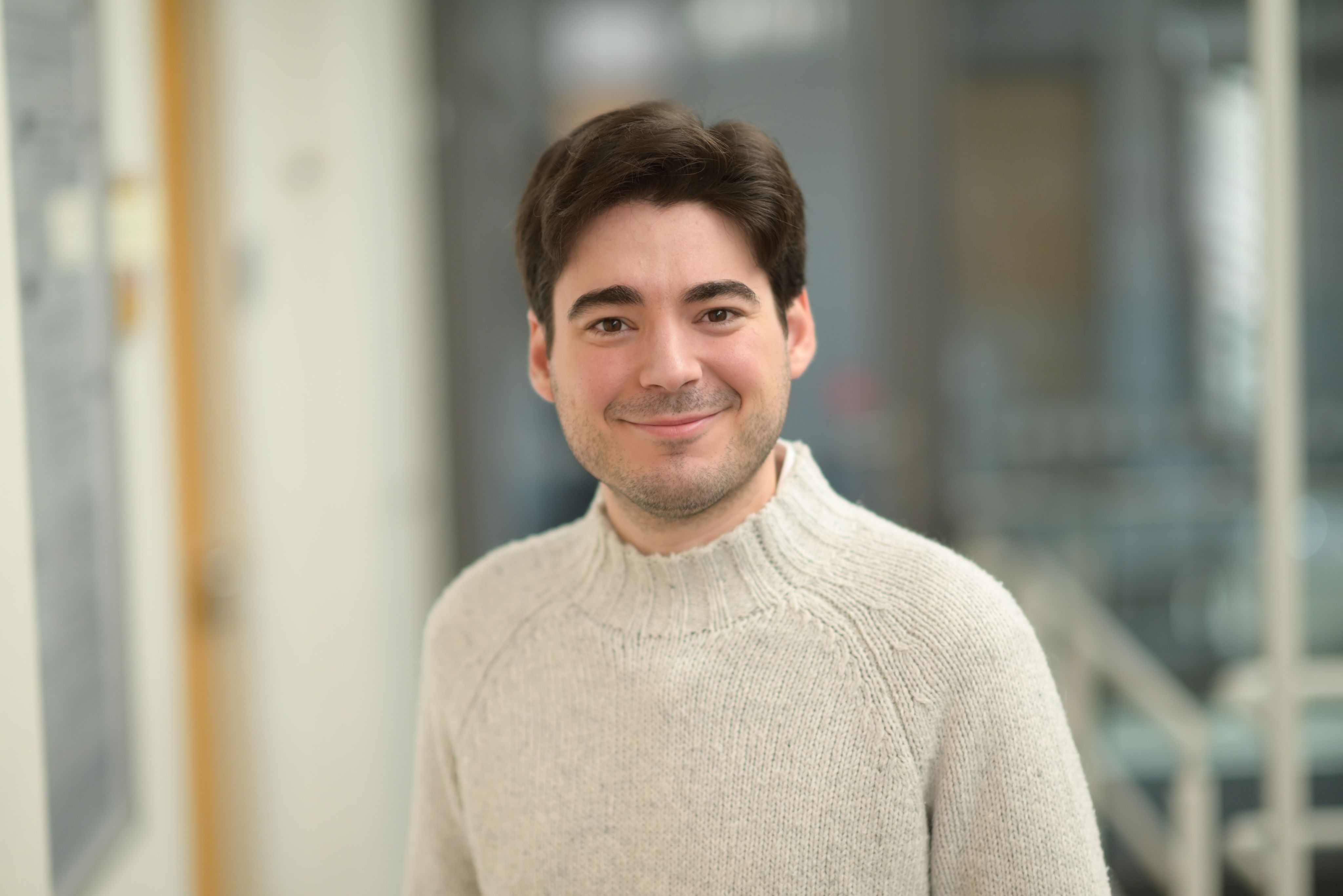 Man with dark hair wearing a beige sweater smiling 