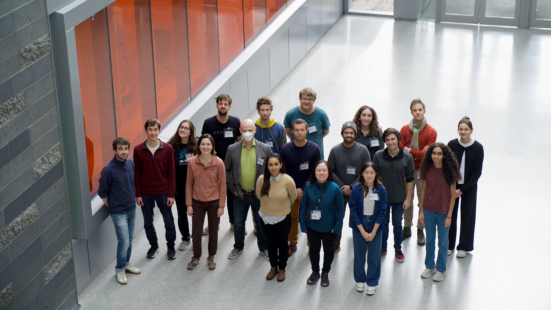 A group of 18 people standing in front of an orange wall in a big hallway looking up
