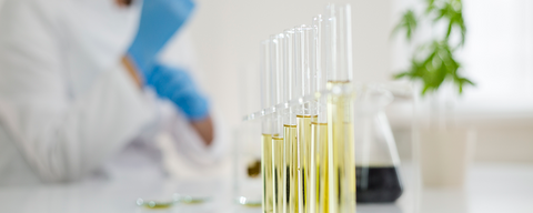 Test tubes with yellow liquid and a person in a white lab coat and blue gloves is in the background.