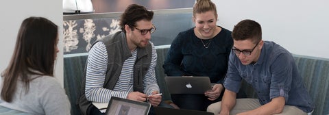 Four grad students with laptops conversing on couches in lounge area.