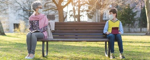 Young and older persons with masks on park bench, looking at each other 