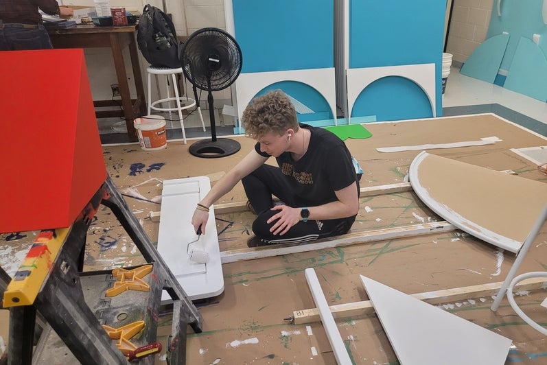 a person sitting on the floor uses a smaller paint roller to paint some wood white