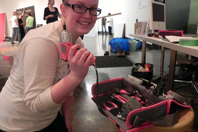 Melissa poses with a small pink drill in hand infront of a full pink tool set in the gallery.