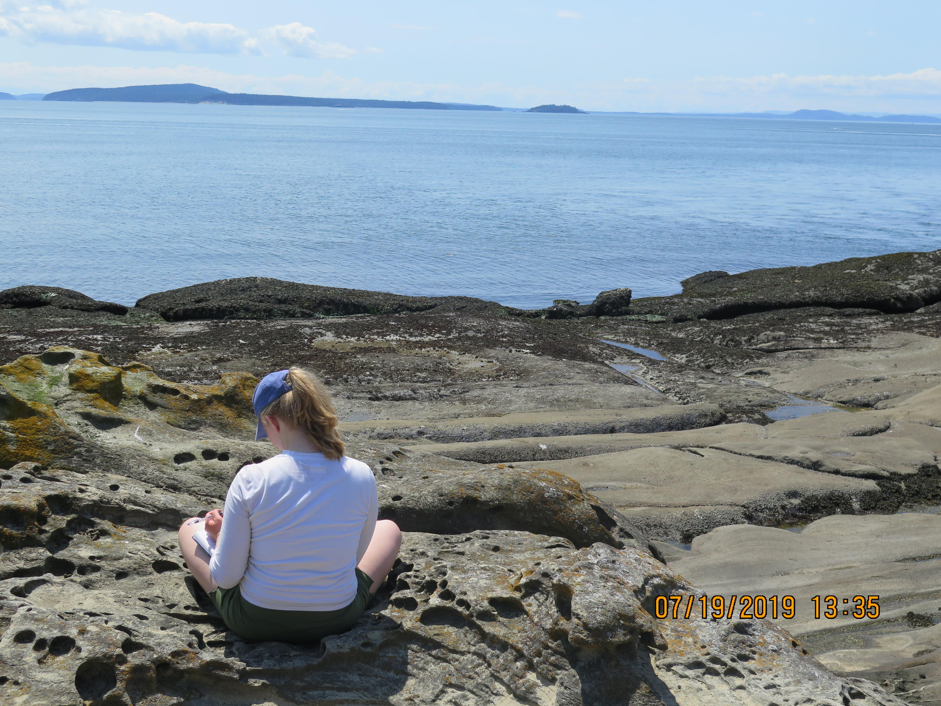 student sitting on rocky shore writing in journal