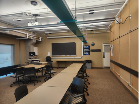 Classroom-style study room with tables, rolling chairs, a large chalkboard, and ceiling-mounted projector.