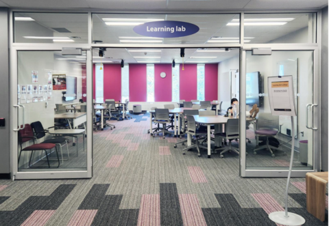 Glass-walled learning lab with tables and chairs, pink accent wall, wall clock, and a person seated inside.