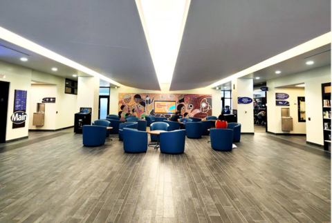 Library lobby with blue lounge chairs arranged around small tables, people seated, and a colourful mural on the back wall.
