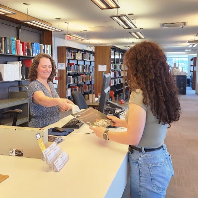 Inside a library, a circulation desk where a librarian hands a book to a customer; shelves of books line the background under bright ceiling lights.