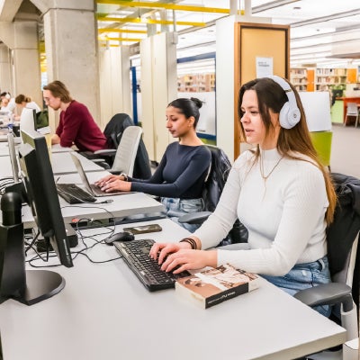 Public study area with several people at computers. Foreground shows a person with long hair wearing headphones and a white top typing on a keyboard