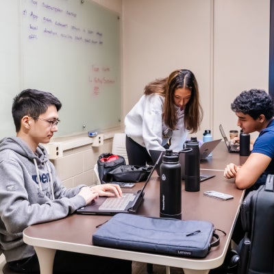 Four students are seated around a table in a classroom, working on laptops.