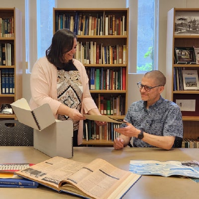 A library scene: a standing woman hands a document to a seated man in a blue patterned shirt at a table with an open book and folders.