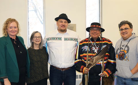Five individuals standing together indoors, wearing a mix of formal and traditional clothing. One person holds a feather.
