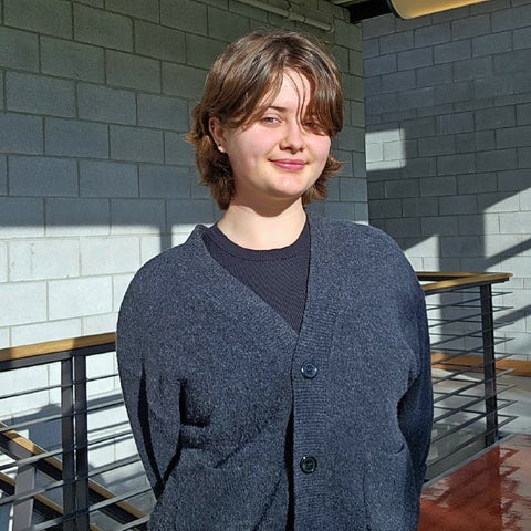 A person with short brown hair wearing a dark cardigan stands in a well-lit space with gray walls and a wooden railing.
