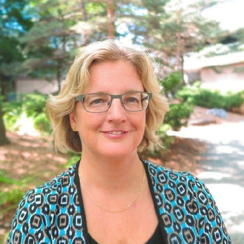 A woman with blonde, shoulder-length hair is wearing glasses and a patterned cardigan. She stands outdoors, surrounded by trees.