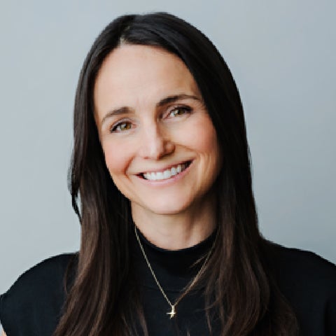 A woman with long dark hair wearing a black top and a gold necklace, standing against a light gray background.