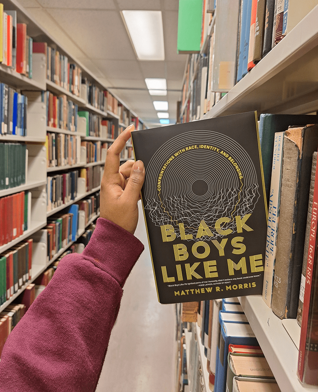 a hand holding the book "Black Boys Like Me" by Matthew R. Morris in a library aisle