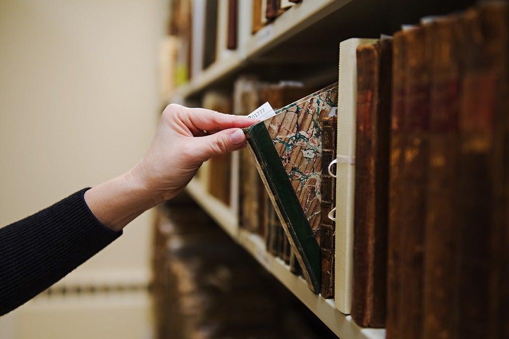 A hand pulling a historical book from an archive collection on a wooden library shelf
