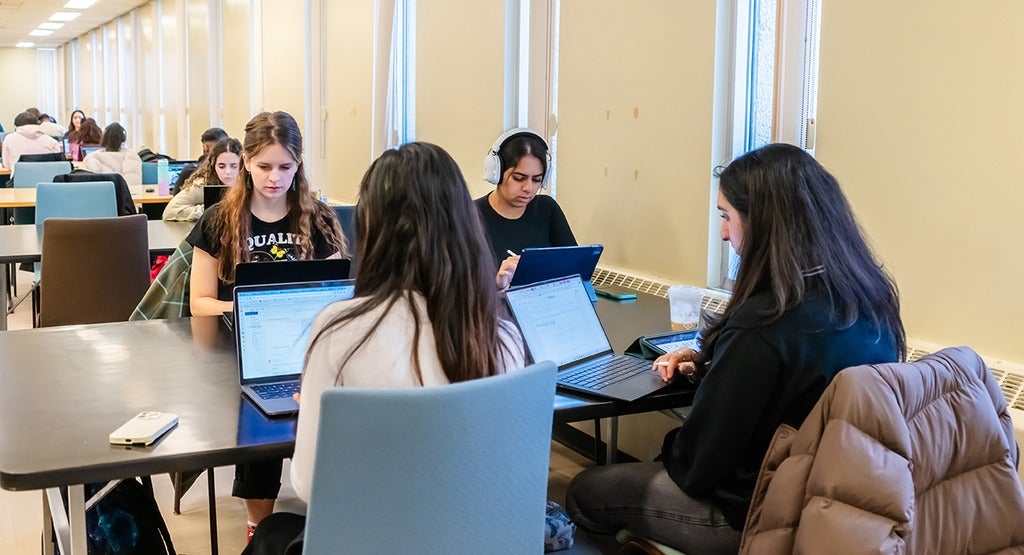 A group of four students sitting at a desk in a library, two with their backs to the viewer. All are working on laptops.