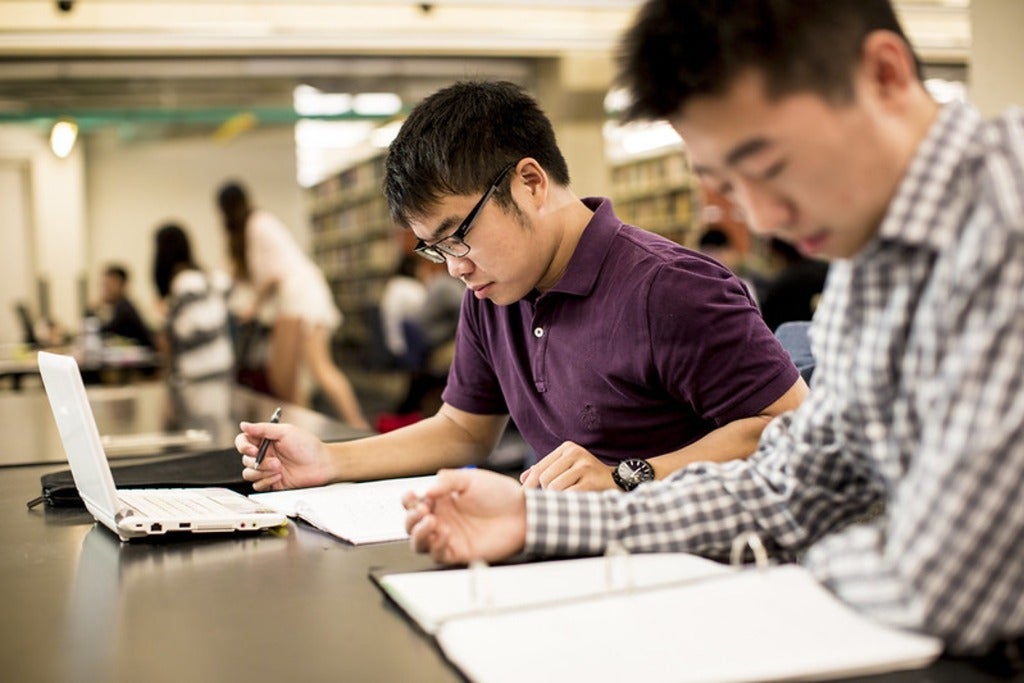 Two students sitting at a table studying, one with a notebook and one with a laptop.