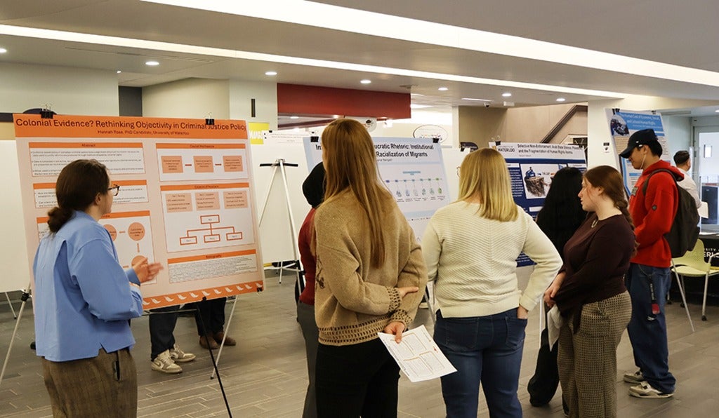 Several people stand in an indoor academic poster session, viewing and discussing research displays mounted on easels