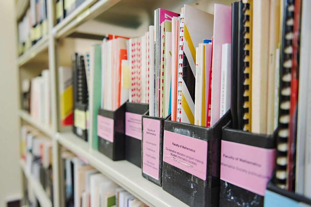 Archival publications and reports organized in labelled document holders on library shelves.