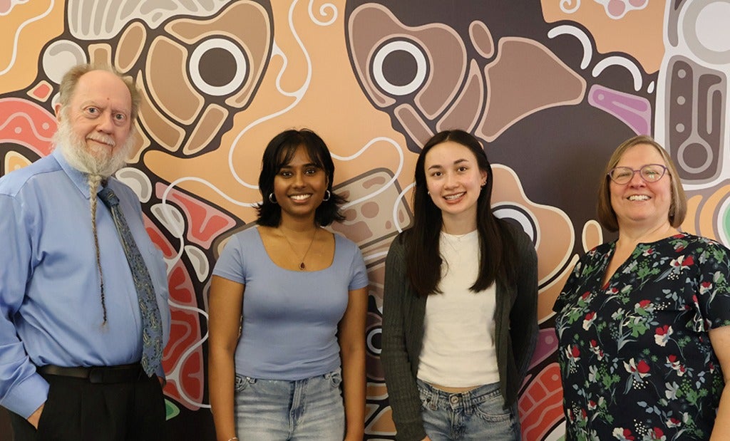 Four people standing in front of a colorful Indigenous-style mural, posing for a group photo indoors