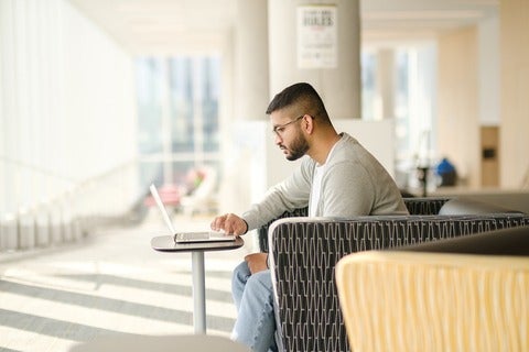 man in a light grey shirt sitting in a chair working on a laptop in an indoor setting