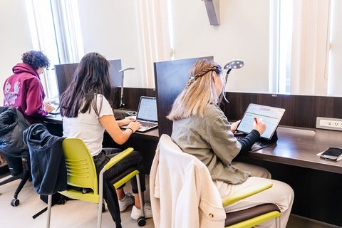 Three students are seated at desks working on laptops in a well-lit study area.