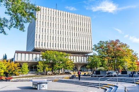 Modern multi-story building with a boxy upper section, in a courtyard with autumn trees and a blue sky.