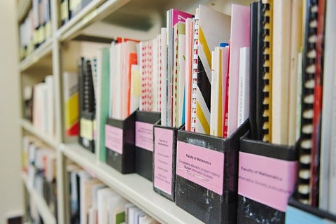 Archival publications and reports organized in labelled document holders on library shelves.