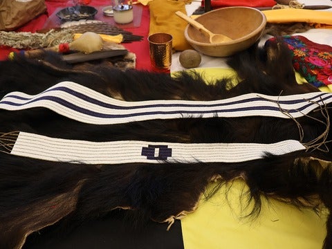 Two woven belts with white and blue patterns laid on a dark fur background, surrounded by various traditional artifacts.